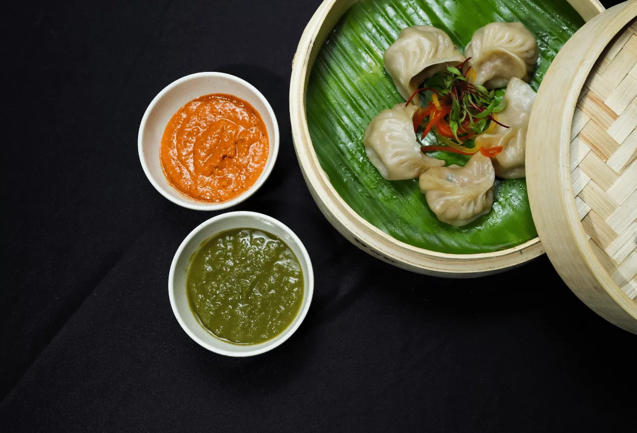 Steamed dumplings with red and green sauces on a green leaf inside a bamboo steamer.