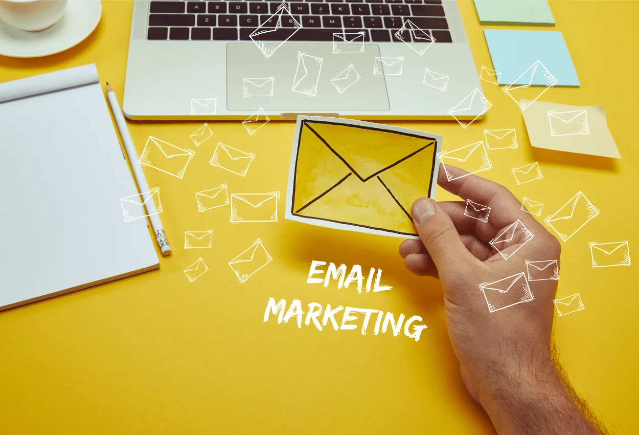 Email marketing concept with laptop, notepad, and hand holding an envelope on a yellow background.