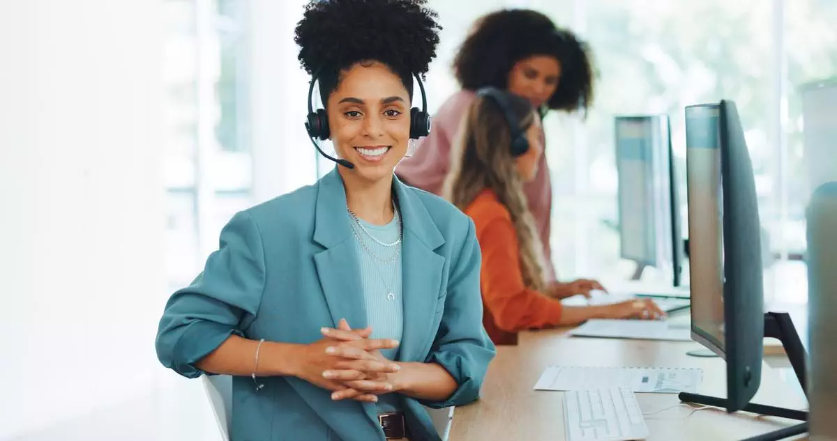 Black woman in call center, wearing headset and smiling, with colleagues working in the background.