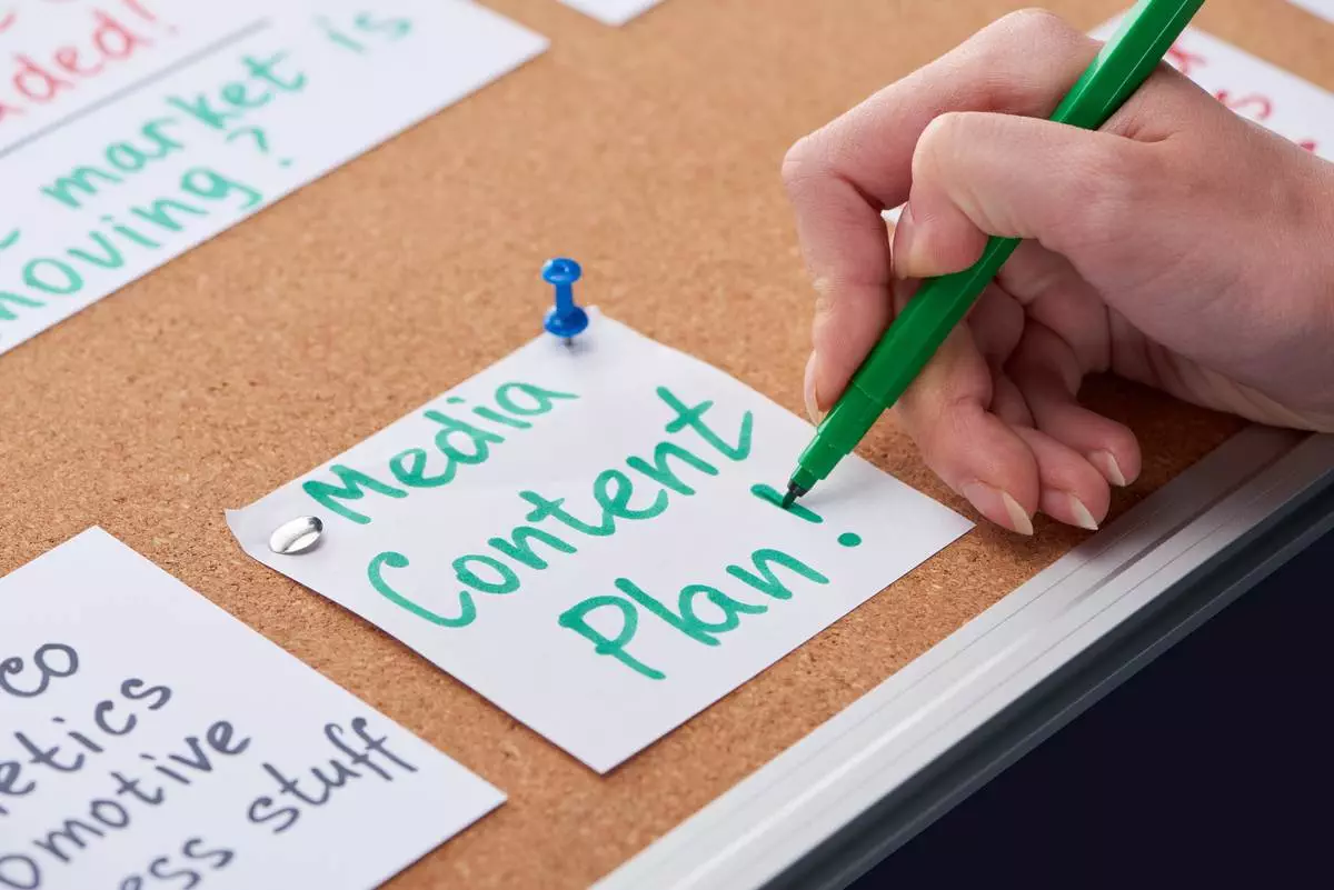 Female hand writing a media content plan on white paper using a green marker, with surrounding notes pinned on a corkboard.