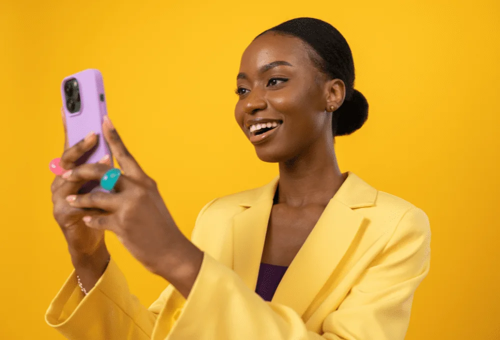 Woman in a yellow blazer taking a selfie with a purple smartphone against a yellow background.