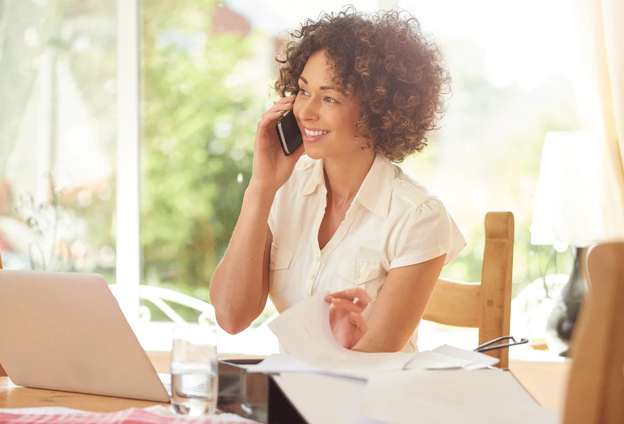 Woman working from home while on a phone call, smiling at her desk with a laptop and paperwork.